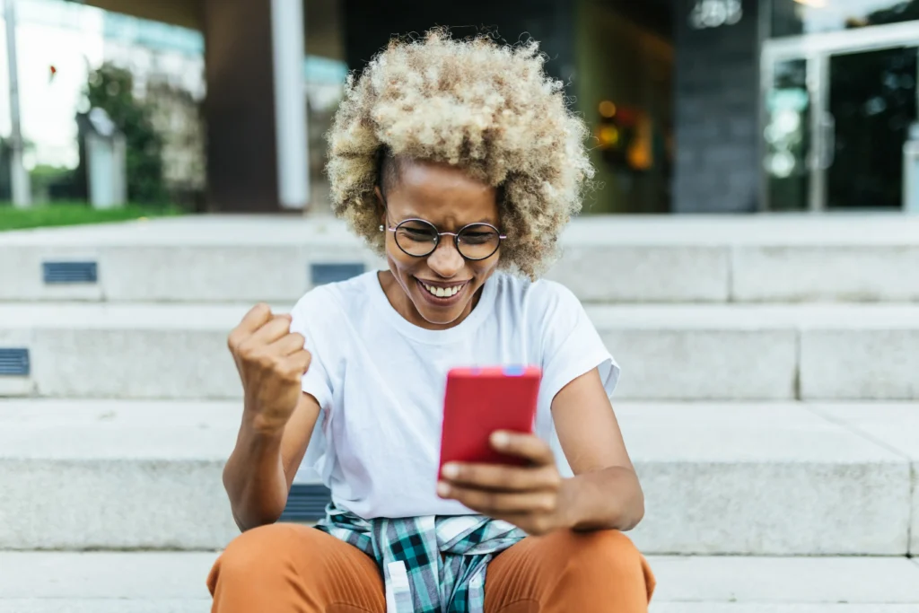 A person with curly hair and glasses is sitting on outdoor steps, looking at a smartphone and raising a fist in excitement.
