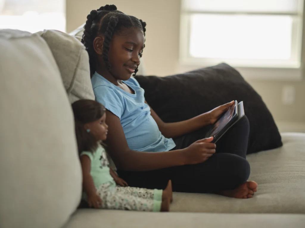 A child, seated on a couch, looks at a tablet while a doll wearing a green outfit sits beside her, symbolizing the importance of representation in toys.