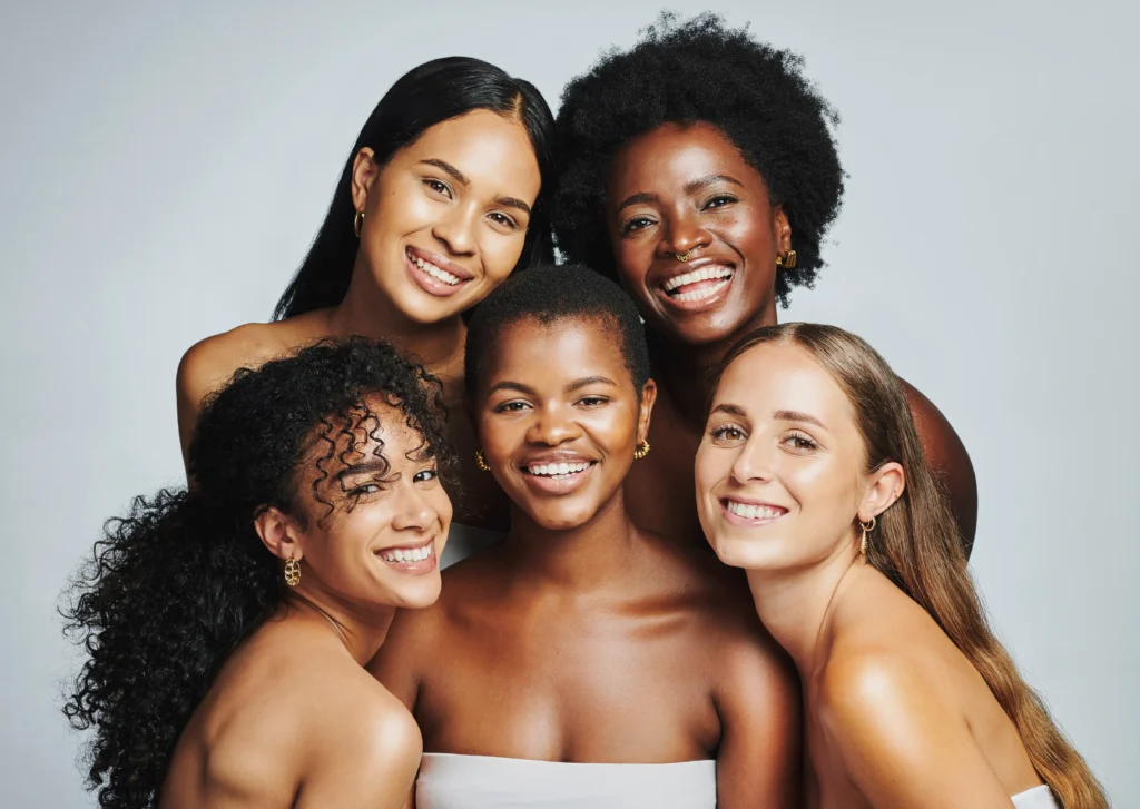 Five women with diverse hairstyles and skin tones are smiling and closely huddled together against a plain background.