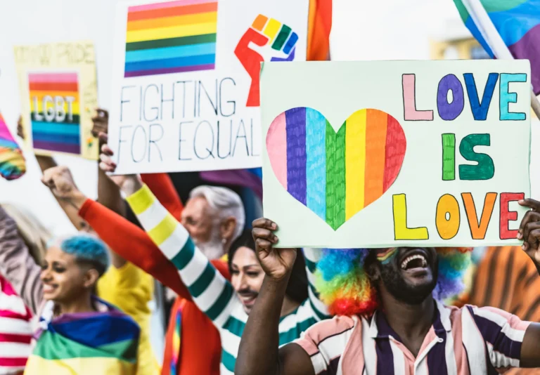 A diverse group of people holding LGBTQ+ pride signs, with messages like "Love is Love" and "Fighting for Equality," participate in a colorful and vibrant pride parade, spreading the message of #OneLove.