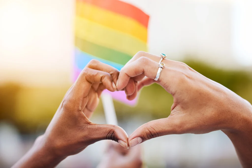 Two hands forming a heart shape in the foreground with a blurred rainbow pride flag in the background, symbolizing unity and love for all. #OneLove