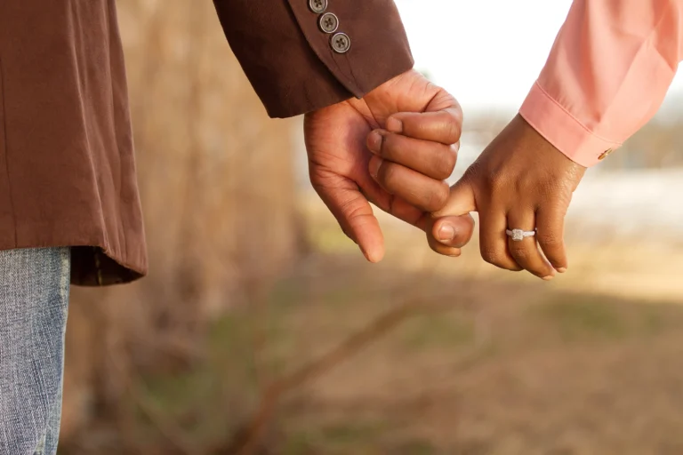 Close-up of two people holding hands, with one wearing a brown jacket and the other a pink shirt adorned with an engagement ring. The background is blurred, emphasizing their connection. #OneLove