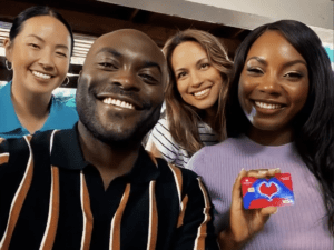 Four people are smiling at the camera, with one person holding a red Visa card showing a heart image. It's an image of the new OneUnited OneLove Debit Card.