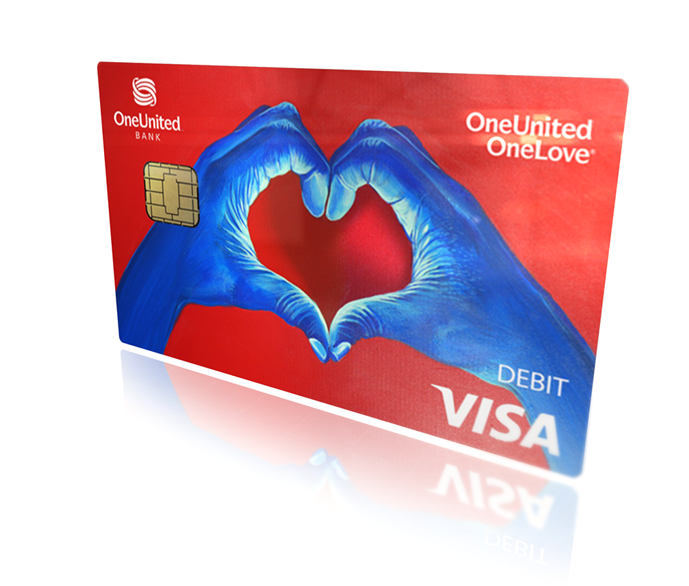 A red OneUnited Bank Visa debit card with blue hands forming a heart shape in the center. The card reads "OneUnited OneLove" at the top and "DEBIT VISA" at the bottom right. Get the OneLove™ Card: Love is where you want to be!