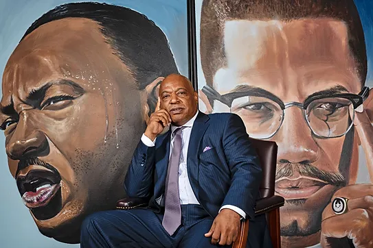 A man in a blue suit sits in a chair between two large portraits, one of Martin Luther King Jr. and the other of Malcolm X.