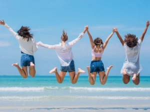 Four people, embodying the essence of financial literacy, jump in unison while holding hands on a beach, facing the ocean with their backs to the camera. They wear casual summer clothing, including shorts and light tops.