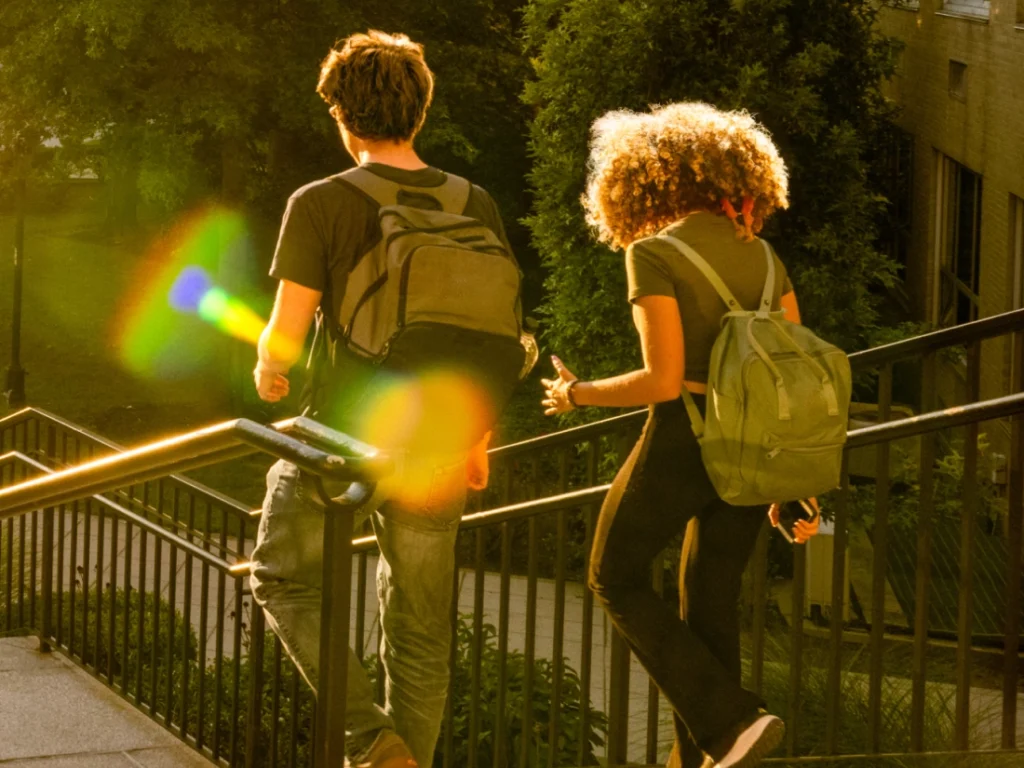 Two people with backpacks walk up an outdoor staircase in the evening sunlight, surrounded by greenery.