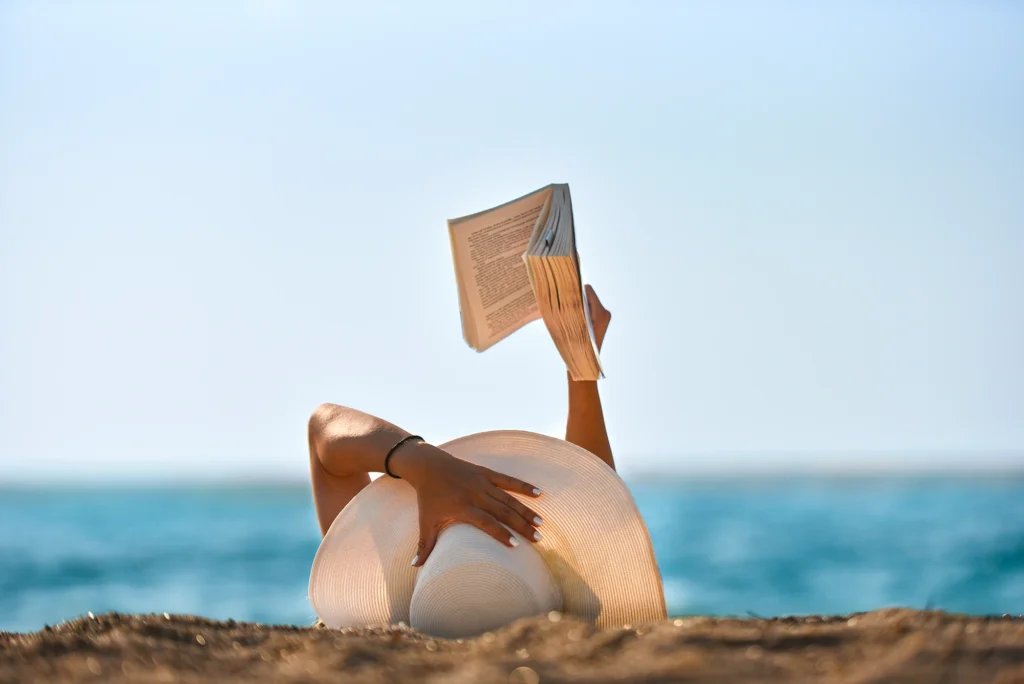 A person lies on a beach with a sun hat, holding an open book up to read about financial love languages. The ocean glistens in the background, framed by a clear day's blue sky.