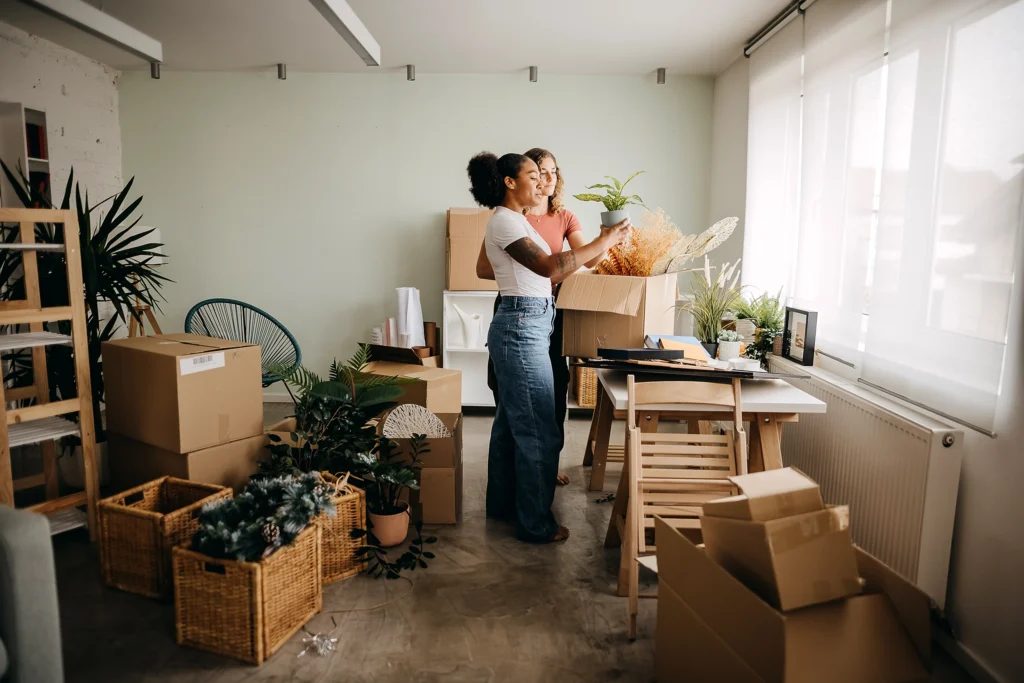 Two people stand in a room surrounded by moving boxes and houseplants, organizing items and unpacking. The room has light-colored walls, large windows, and several scattered pieces of furniture.