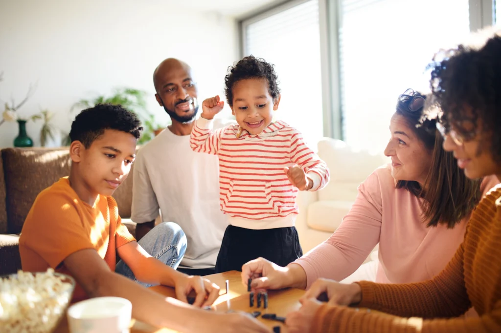 A family of five is gathered around a table playing a financial literacy game in a brightly lit room. The young child in the center is standing and looks excited.