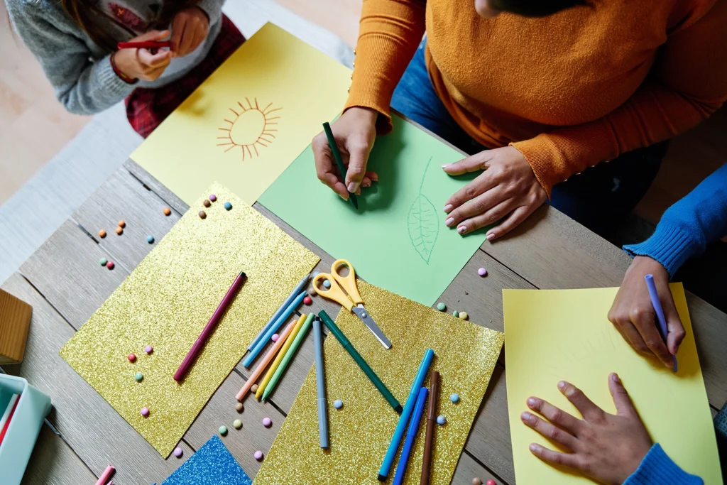 Three people are drawing on colored paper at a table with scattered art supplies, including colored pencils, markers, scissors, and glitter paper. As they create their artwork, they discuss financial literacy and ways to creatively manage their budgets.