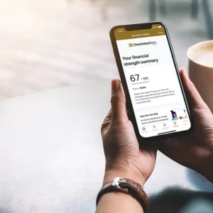 Person holding a smartphone displaying a financial strength summary with a score of 67 out of 100 from OneUnited Bank. An AI-powered personal finance tool behind the scenes makes it easy to track. A cup of coffee is on the table in the background.