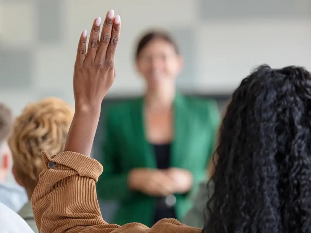 A person with long curly hair raises their hand in a classroom while a teacher in a green blazer, who teaches financial literacy, stands in the background.