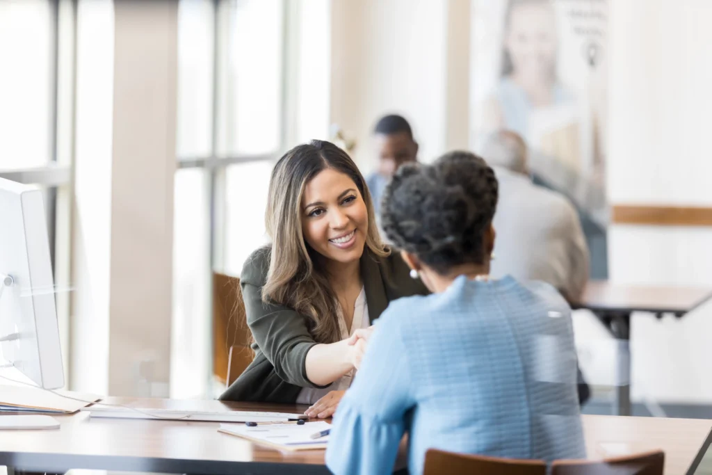 Two women are seated across from each other at a table in a bright, open office. One woman is smiling and extending her hand for a handshake, while the other woman is facing her, listening attentively.