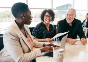 Three colleagues seated at a table smile and engage in a lively conversation, with documents and a laptop open in front of them.