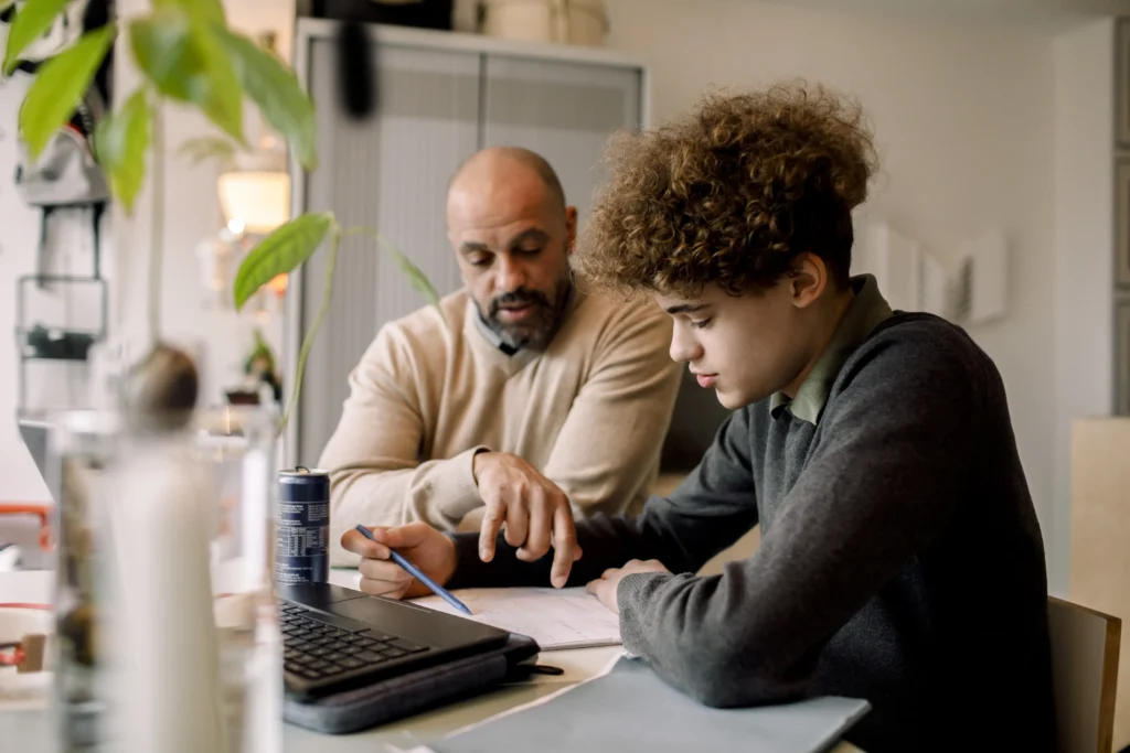 An adult and a teenager sit at a table, focusing on a financial document. The adult points at the paper while the teenager listens, with a laptop and beverage can in the foreground.