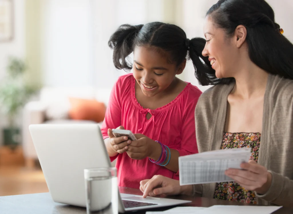 A woman and a girl with ponytails sit at a table. The woman is holding a piece of paper and using a laptop, while the girl looks at a smartphone. A glass of water is also on the table.