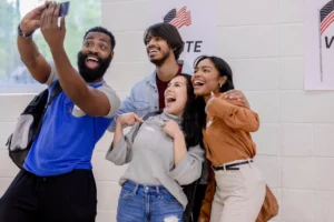 A group of four people smile while taking a selfie in front of a "Vote" sign at a polling location.