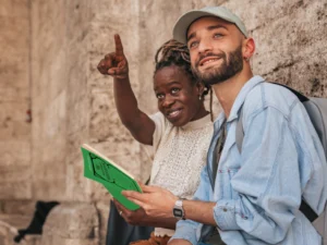 Two people sit against a stone wall; one is holding a green book and smiling, while the other is pointing towards something in the distance.