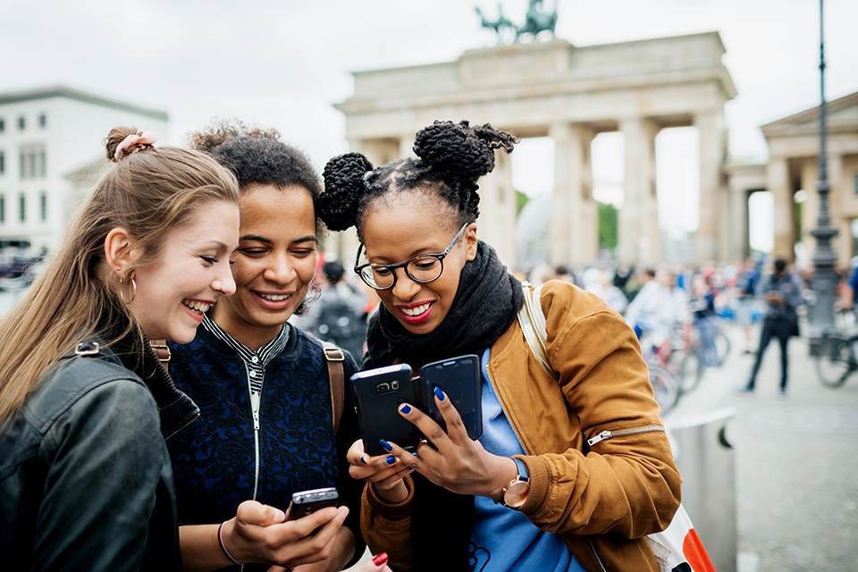 Three people are standing in front of the Brandenburg Gate, smiling and looking at their phones, capturing memorable moments on their Black Travel adventure.