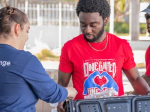 A man in a red shirt and a woman in a blue shirt are sorting items at an outdoor event. The man holds a container, and the woman places items inside it.