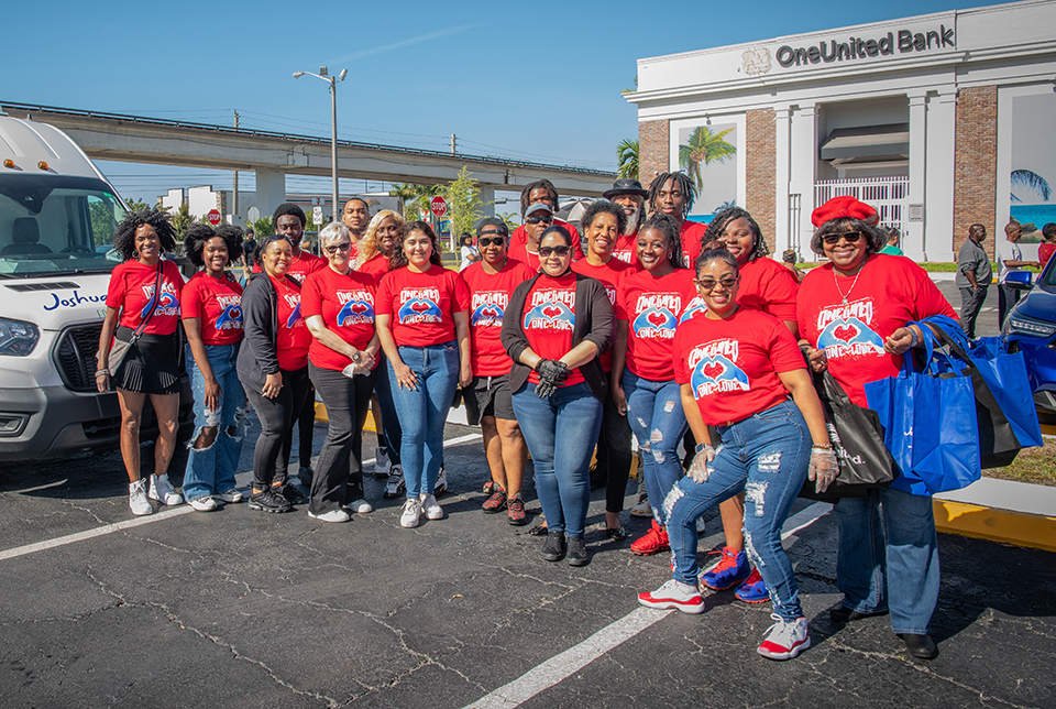 A group of people wearing red "Unity" shirts pose for a photo in a parking lot in front of community bank, OneUnited Bank.