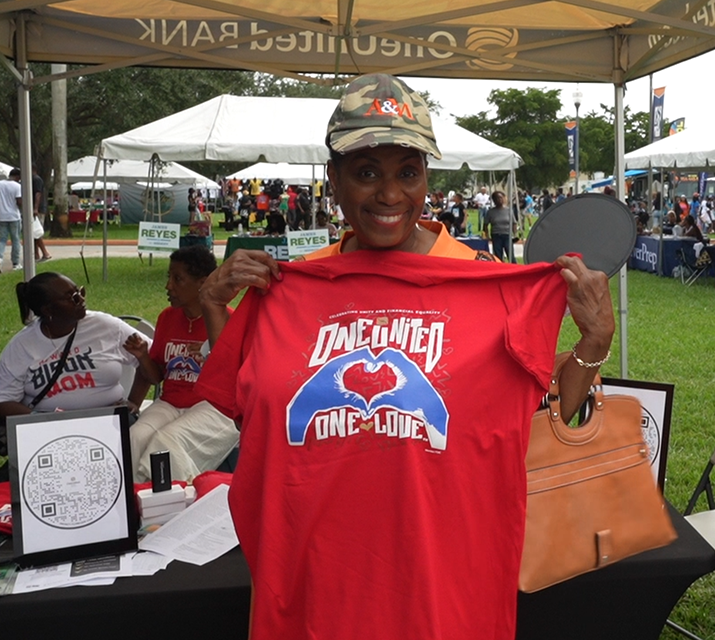 A person stands under a tent at an outdoor event, smiling and holding up a red T-shirt that reads "One Love One United." Various booths and people, including representatives from community banks, are visible in the background.