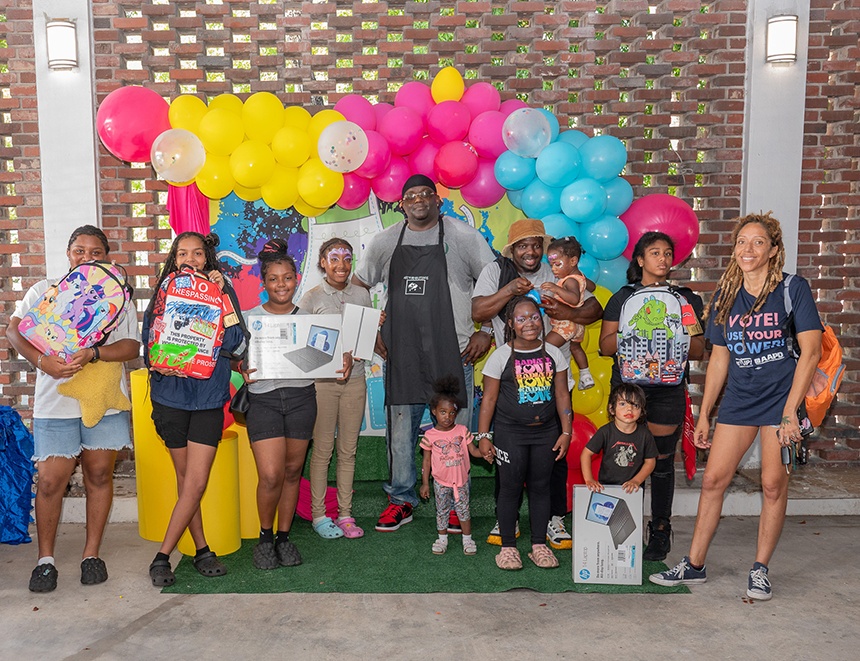 A group of people, including children, pose with backpacks and boxes in front of colorful balloons and decorations, showcasing the support from community banks.