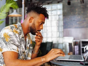 A man with short curly hair and a beard wearing a patterned shirt works intently on a laptop at a table in a modern, industrial-style setting.