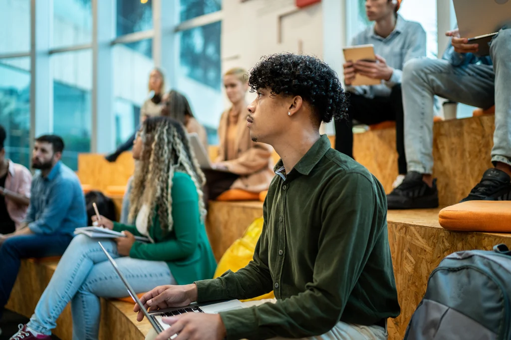 A diverse group of people sitting on tiered wooden benches in a modern, well-lit room, some using laptops and notepads.