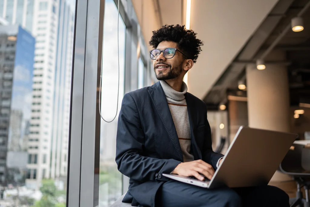 A person with glasses and curly hair sits by a window using a laptop, looking outside and smiling. They are dressed in a dark blazer and a light turtleneck sweater. Urban buildings are visible outside.