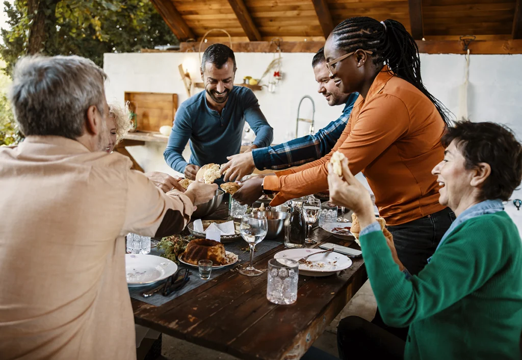 A group of people sit around a wooden table outdoors, sharing food and laughing, while passing a dish to each other, their bonds strengthened by love and the joy of simple pleasures over wealth.