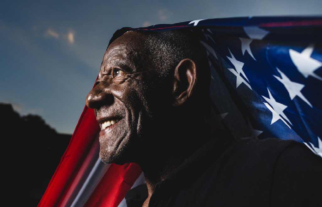 An older man smiles while draped in an American flag, with a dark sky in the background.