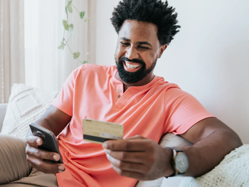 Man in a coral shirt smiles while holding a smartphone and a credit card, sitting on a couch.