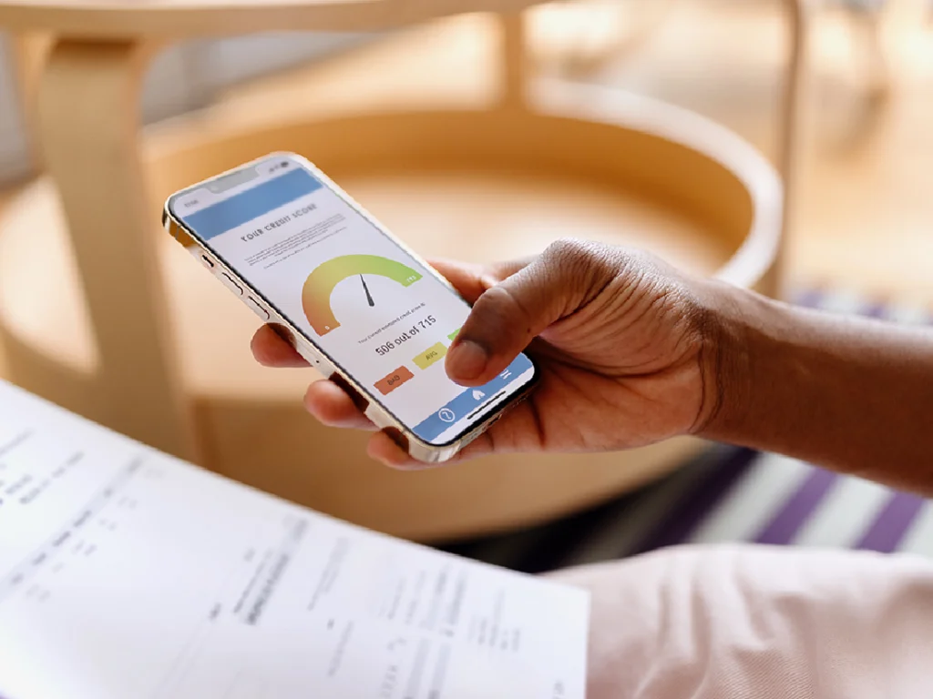 Person holding a smartphone displaying a credit score gauge with numerical score; documents are visible in the background.