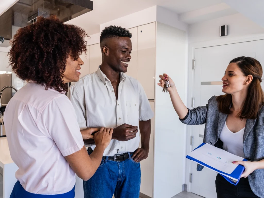 A real estate agent hands keys to a smiling couple, thrilled by their new modern home, thanks to the First-Time Homebuyer Assistance they received.