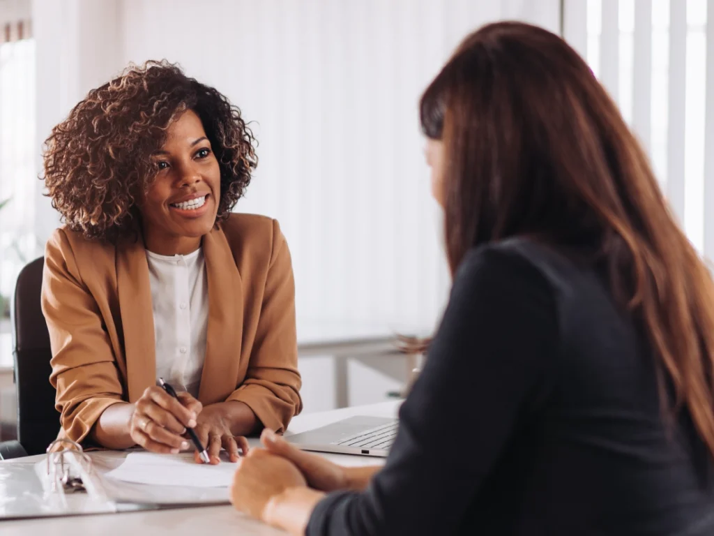 Two people sit at a desk in an office setting, one smiling and holding a pen, possibly discussing or signing documents related to First-Time Homebuyer Assistance.