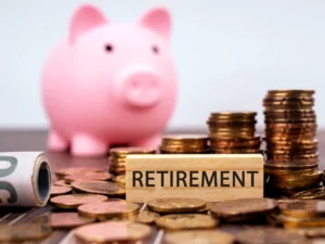 A pink piggy bank and stacks of coins artfully surround a small wooden block labeled "RETIREMENT," symbolizing careful planning for one's golden years.