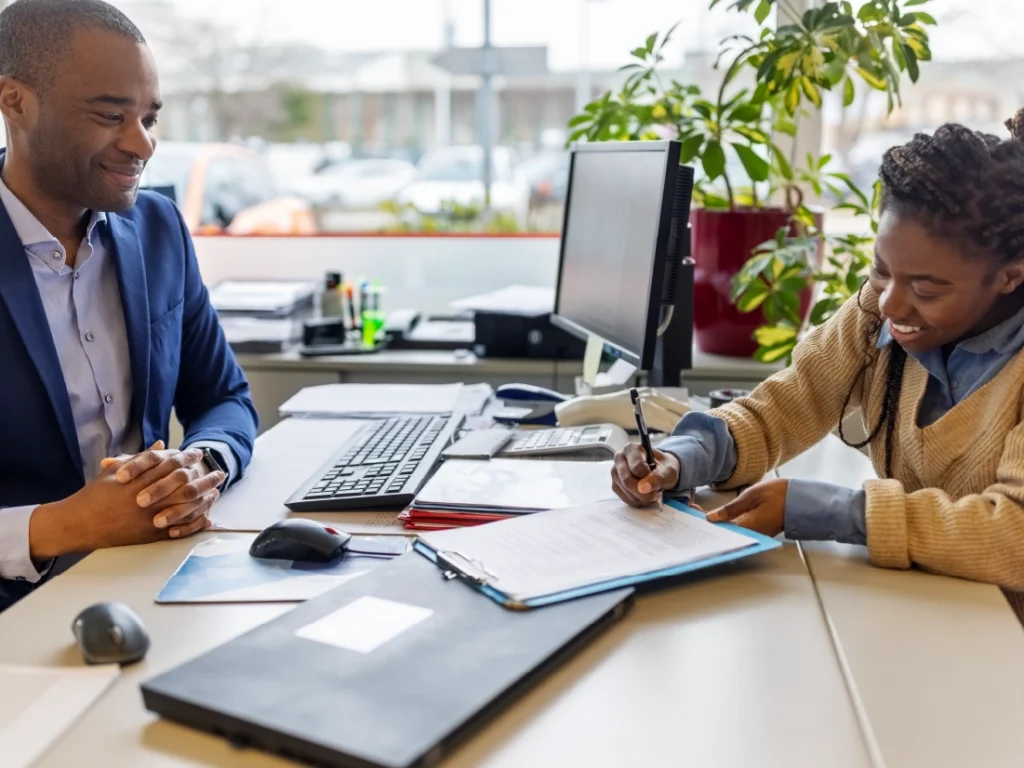 A woman signs retirement documents at a desk while a man sits across from her. They are in an office adorned with a computer and plants.