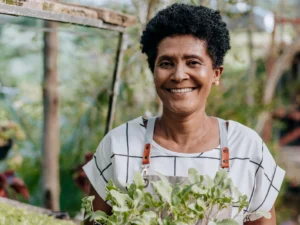 A person smiling while holding a plant outdoors, surrounded by greenery.