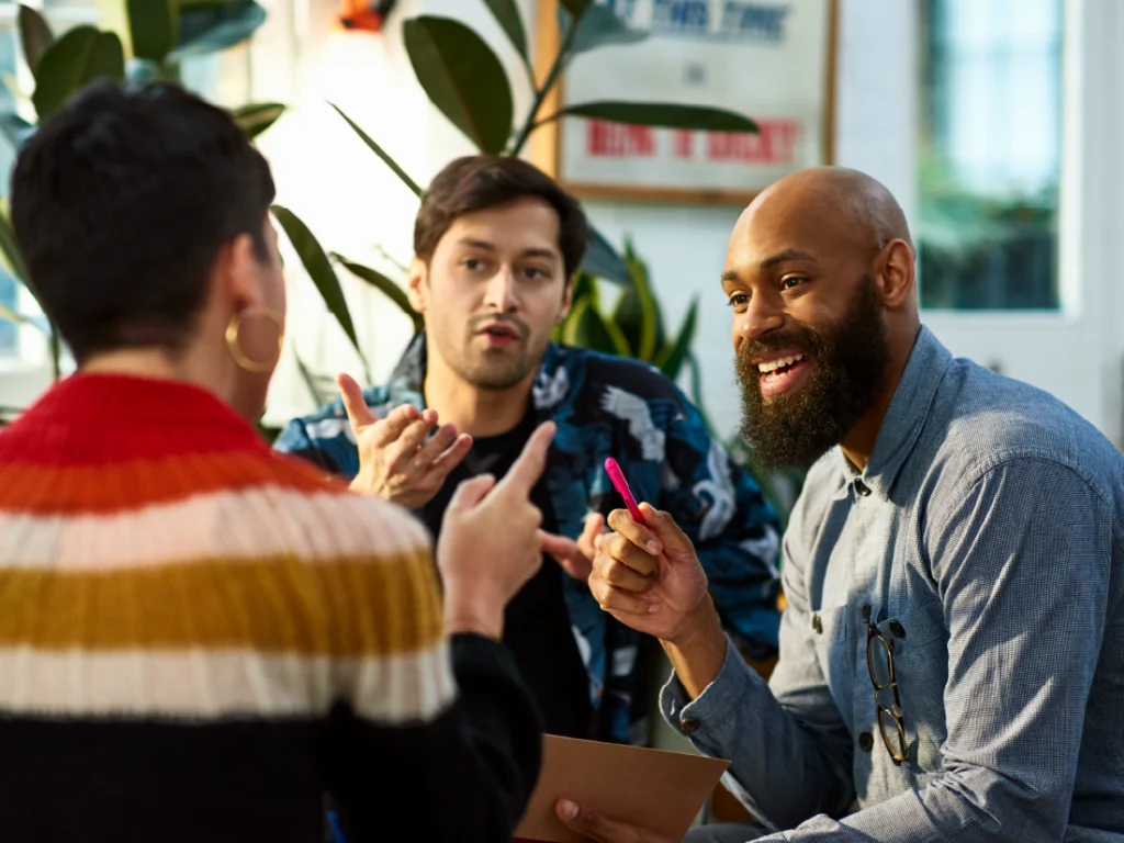 Three people are engaged in a lively discussion in a bright room with plants. One person holds a pen, while another gestures with their hands.