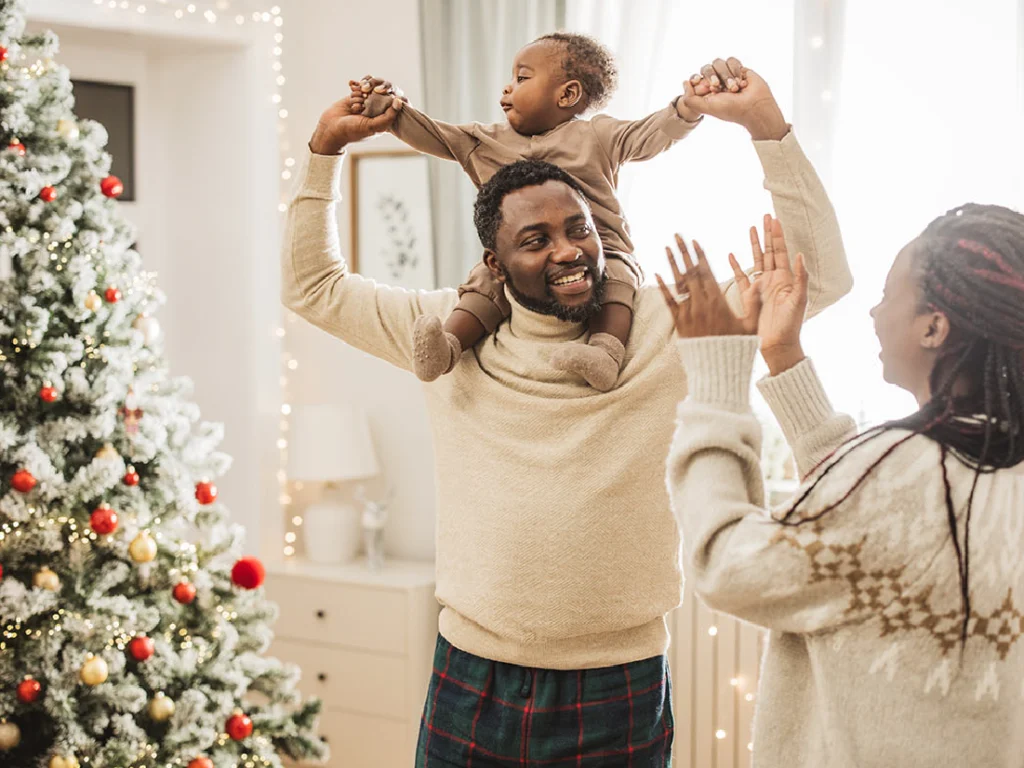 A man holds a child on his shoulders beside a decorated Christmas tree, with a woman clapping nearby in a festive room.