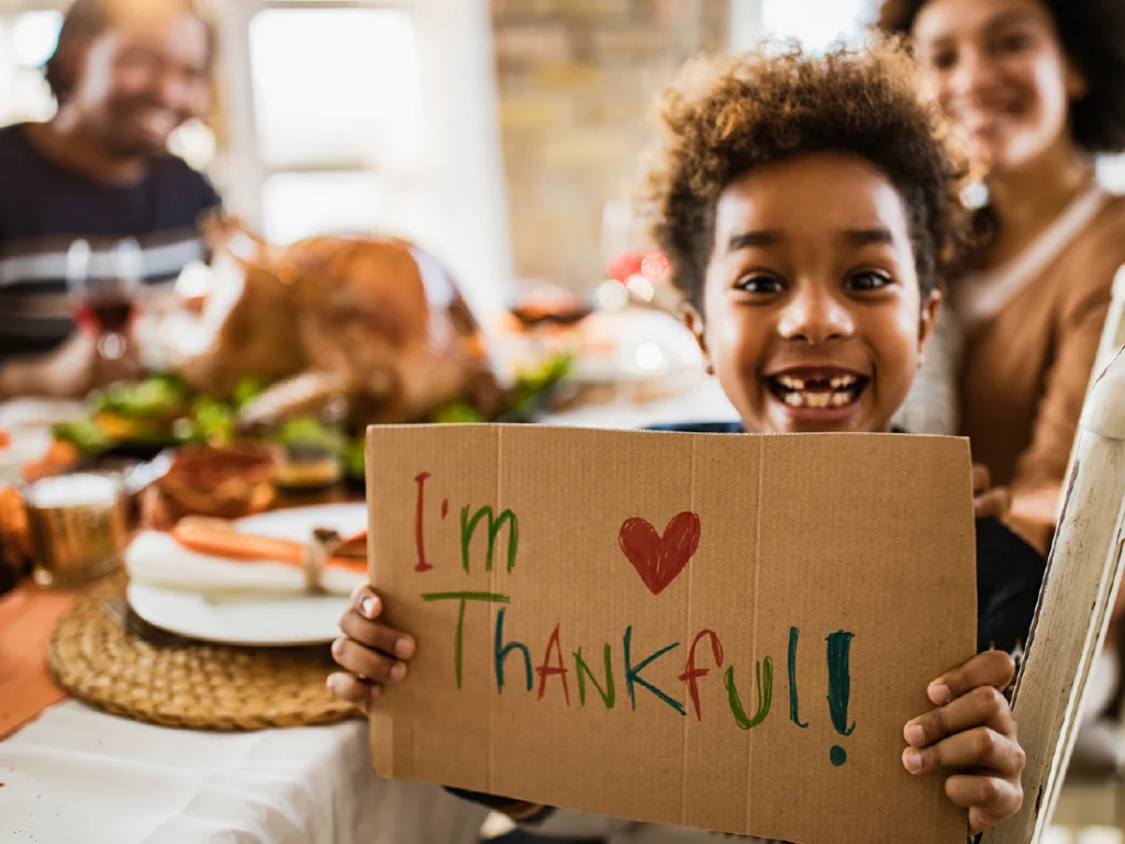 A smiling child holds a sign reading "I'm Thankful!" at a table with a Thanksgiving meal, including a turkey, in the background.