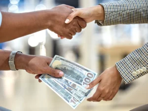 Two individuals shaking hands while engaging in a lending transaction, exchanging several U.S. hundred-dollar bills.