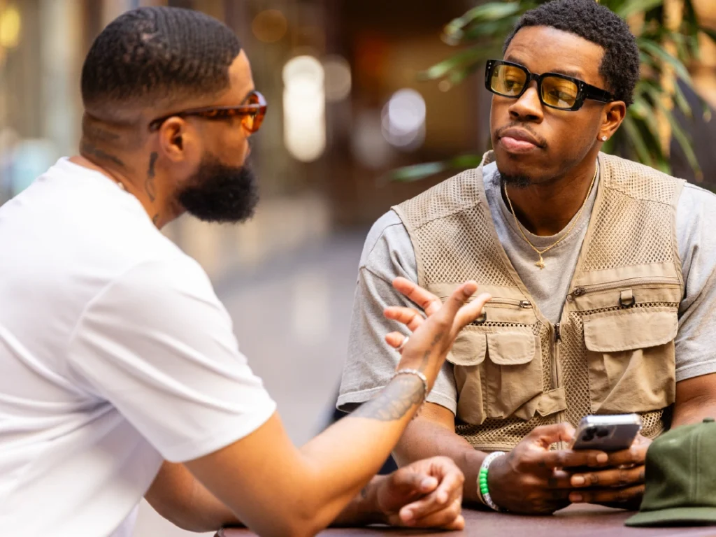 Two men sit at a table in conversation. One man gestures while speaking about lending, and the other listens intently, holding a smartphone. Both wear glasses, immersed in their casual setting.