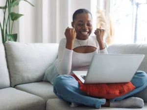 A person sitting cross-legged on a couch is smiling and raising their fists in excitement while looking at a laptop, thrilled by the success of their latest lending venture.