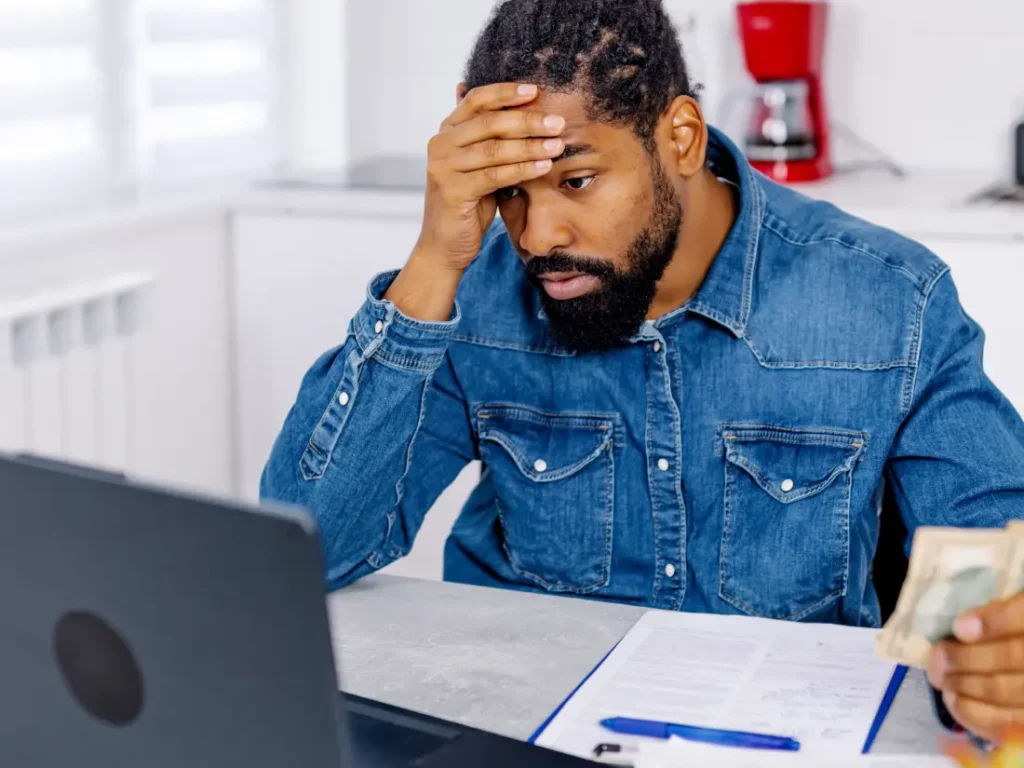 A man in a denim shirt looks worriedly at a laptop while holding cash, sitting at a table surrounded by documents that hint at looming debt.