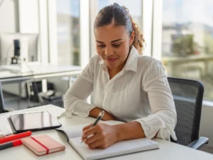 Person in a white shirt writing in a notebook at a desk with a tablet and red notebook nearby. Office setting with large windows in the background.