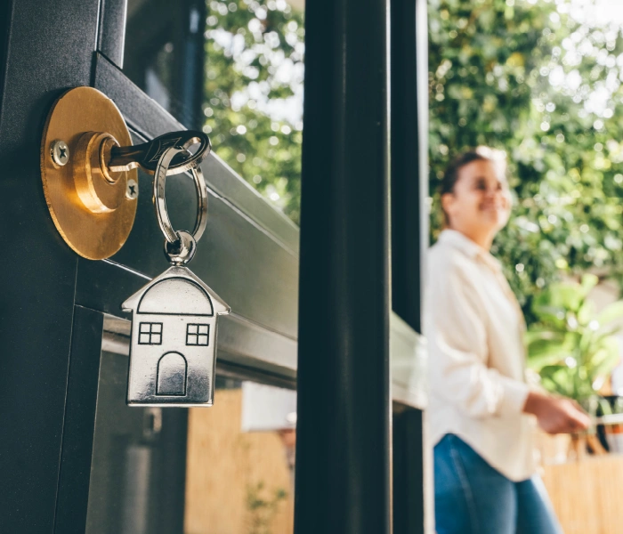 A house-shaped keychain is inserted in a lock on a glass door. A person in casual attire stands in the background.