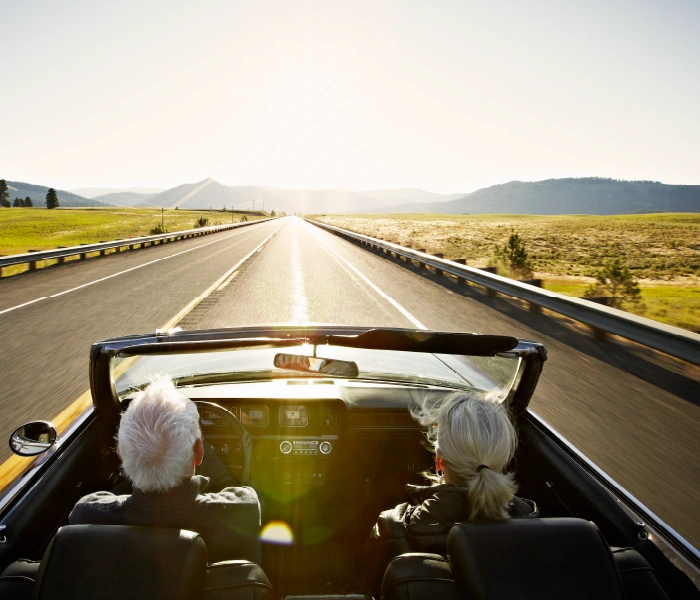 Two people with white hair drive a convertible down an empty highway surrounded by scenic fields and hills under a bright sun.